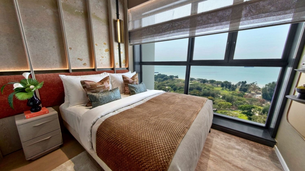 Interior view of a Meyer Blue (Tel:+6587865200) bedroom with a large window overlooking a coastal landscape. A made bed with decorative pillows and a throw blanket is in the foreground, with a bedside table and lamp visible. The window shows a view of the sea, green trees, and a distant shoreline.