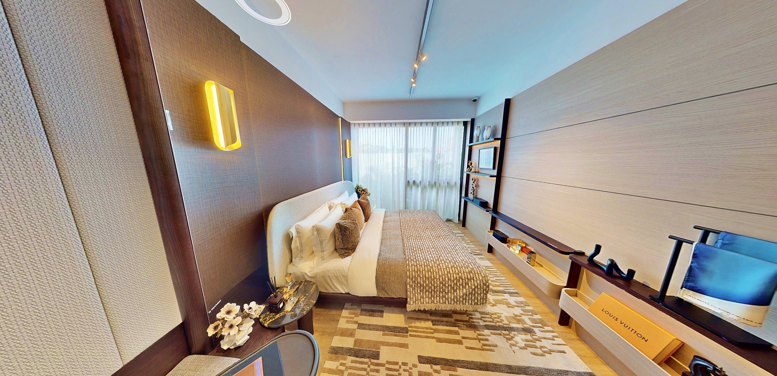Interior photograph of a narrow bedroom with a made bed, built-in shelving, and a "LOUIS VUITTON" box, featuring a minimalist design.