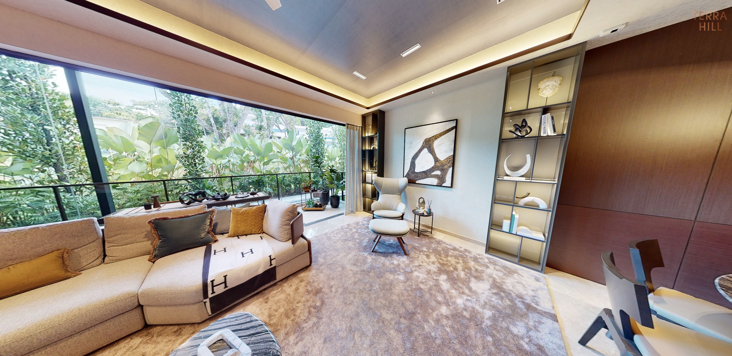 Interior view of a modern living room in Terra Hill condominium, featuring a beige sectional sofa, a large window overlooking greenery, and a built-in shelving unit with decorative items.