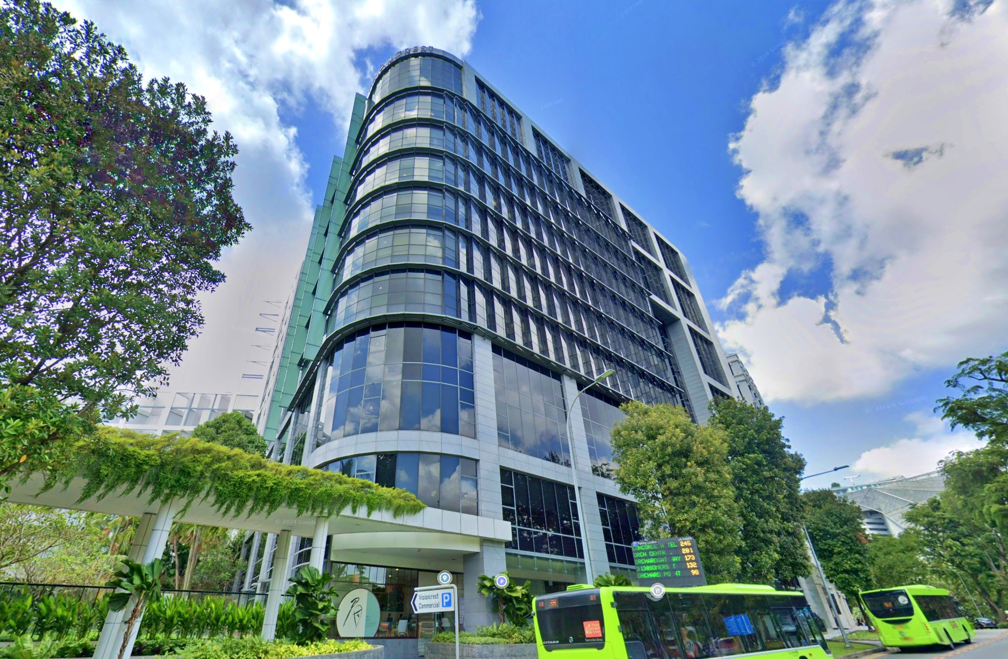 A low-angle shot of the Visioncrest Commercial building in Singapore, featuring its curved facade, glass windows, and greenery at the base, with a bright blue sky and fluffy white clouds above.