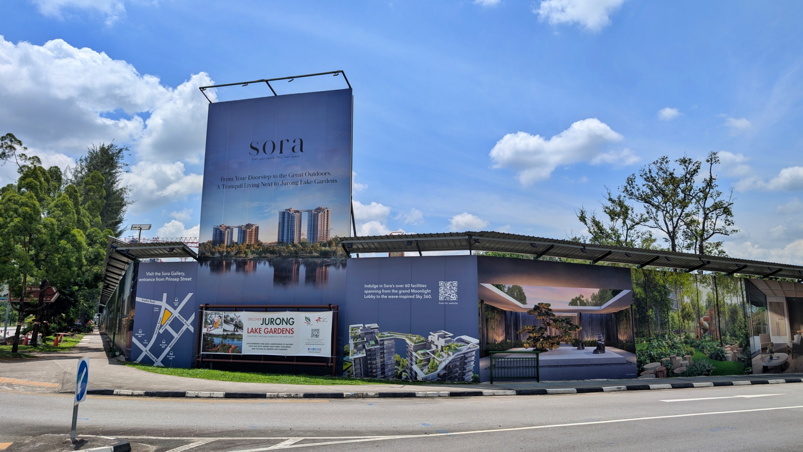 A large, temporary hoarding surrounds a construction site or sales gallery for "sora," a residential development. The hoarding features promotional imagery and text. The tallest section displays the word "sora" in elegant, light grey lettering above an artist's impression of modern apartment buildings reflected in a body of water, possibly a lake or reservoir. Text below this image reads "Find Your Sanctuary in the Lap of Outdoors. Tranquil Living Next to Jurong Lake Gardens." Smaller panels attached to the main hoarding provide additional information, including a map showing the location relative to "Yuan Ching Road," another artist's rendering of the development, a QR code, and the logo for "JURONG LAKE GARDENS." The hoarding is situated along a road with a pedestrian walkway and a blue and white circular sign. Lush green trees are visible in the background under a partly cloudy blue sky.