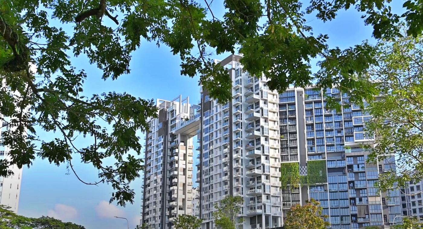 A view of three modern, high-rise residential buildings in Singapore, partially framed by lush green tree branches in the foreground. The buildings feature a contemporary design with numerous balconies and large windows, suggesting apartment or condominium units. The central building is particularly distinctive, with a sky bridge connecting two separate towers at a high level. The buildings are painted in light colors with contrasting darker accents. The sky above is bright blue with a few scattered white clouds. The surrounding area appears to be a mix of urban development and green spaces, with the tops of more trees visible below the buildings.