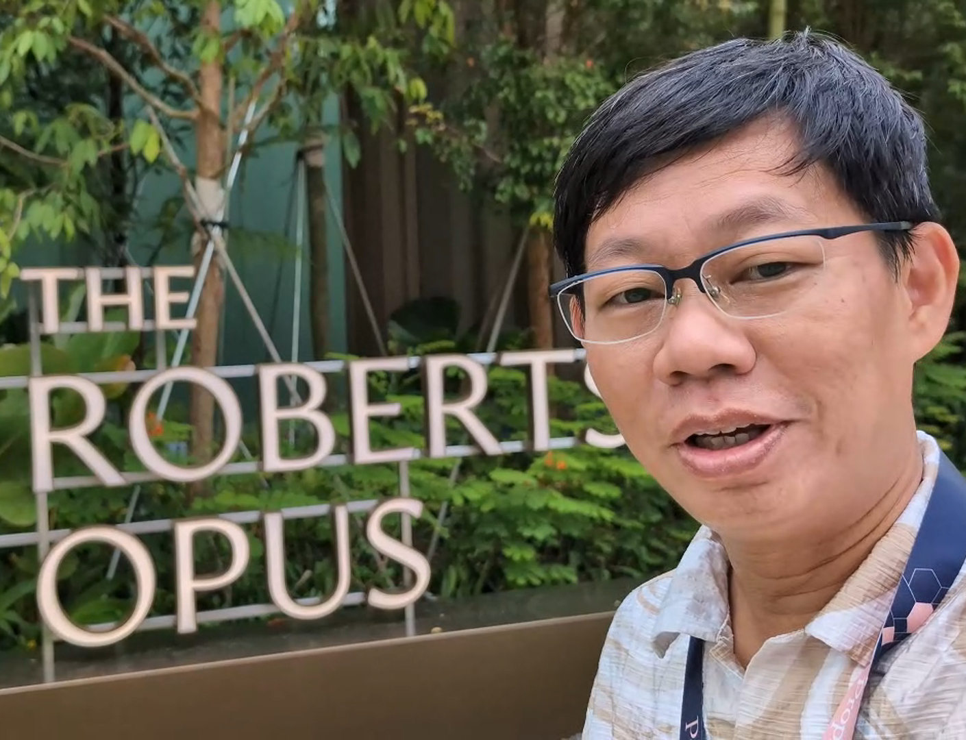 A man with dark hair and glasses smiles slightly, standing in front of a sign that reads "THE ROBERTSON OPUS" and has a decorative "R" logo. Lush green foliage is visible in the