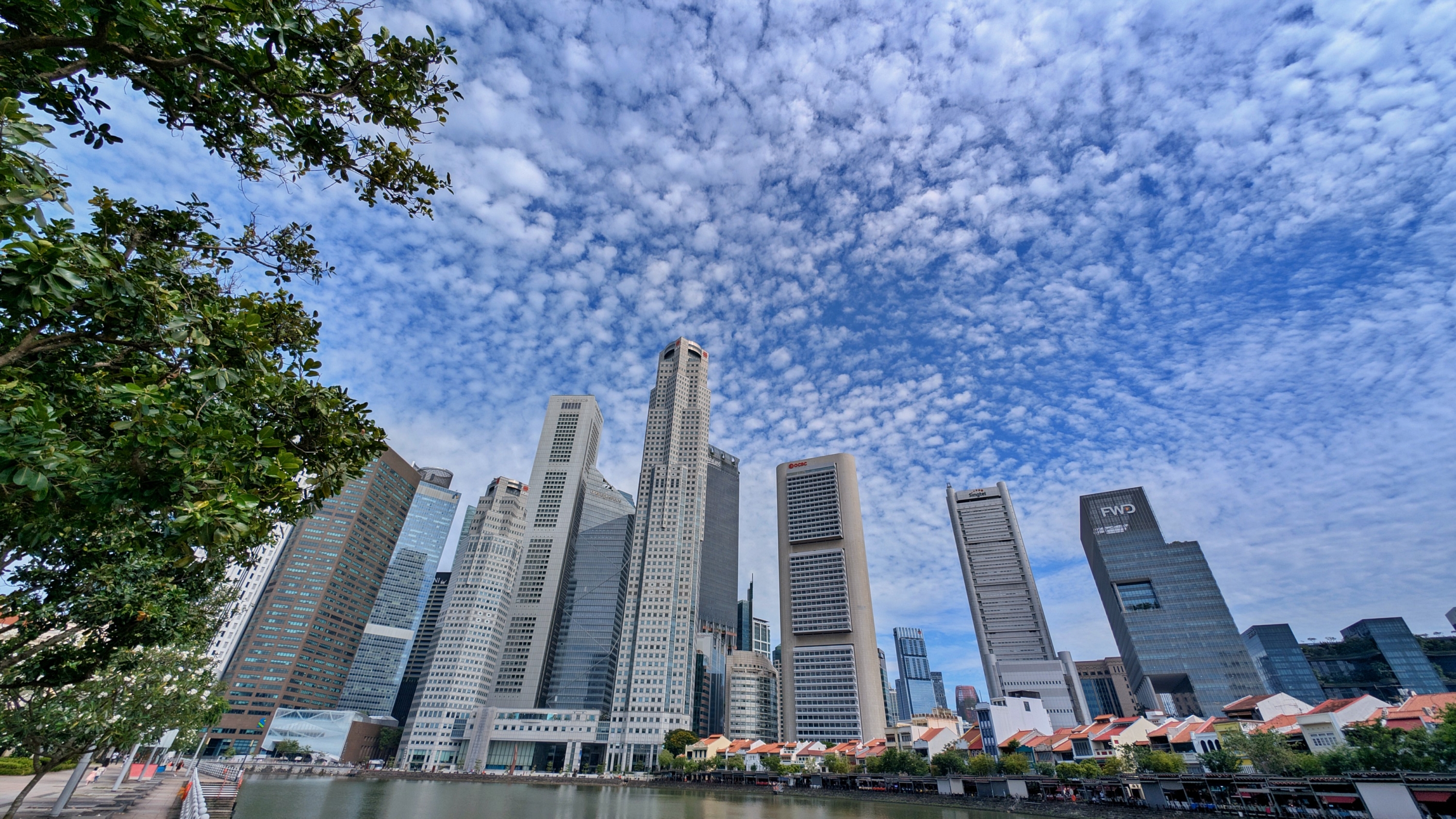 A wide-angle view of the Singapore CBD skyline from the river level, showing numerous modern skyscrapers against a textured cloudy sky.