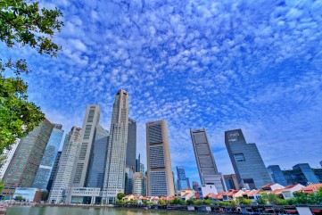 A wide-angle view of the Singapore CBD skyline from the river level, showing numerous modern skyscrapers against a textured cloudy sky.