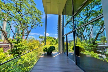 A photograph of a modern balcony with a wooden floor and a glass railing, looking out onto a lush green landscape of trees and buildings, with the clear blue sky above. The reflection of the trees is visible on the large glass windows of the building.