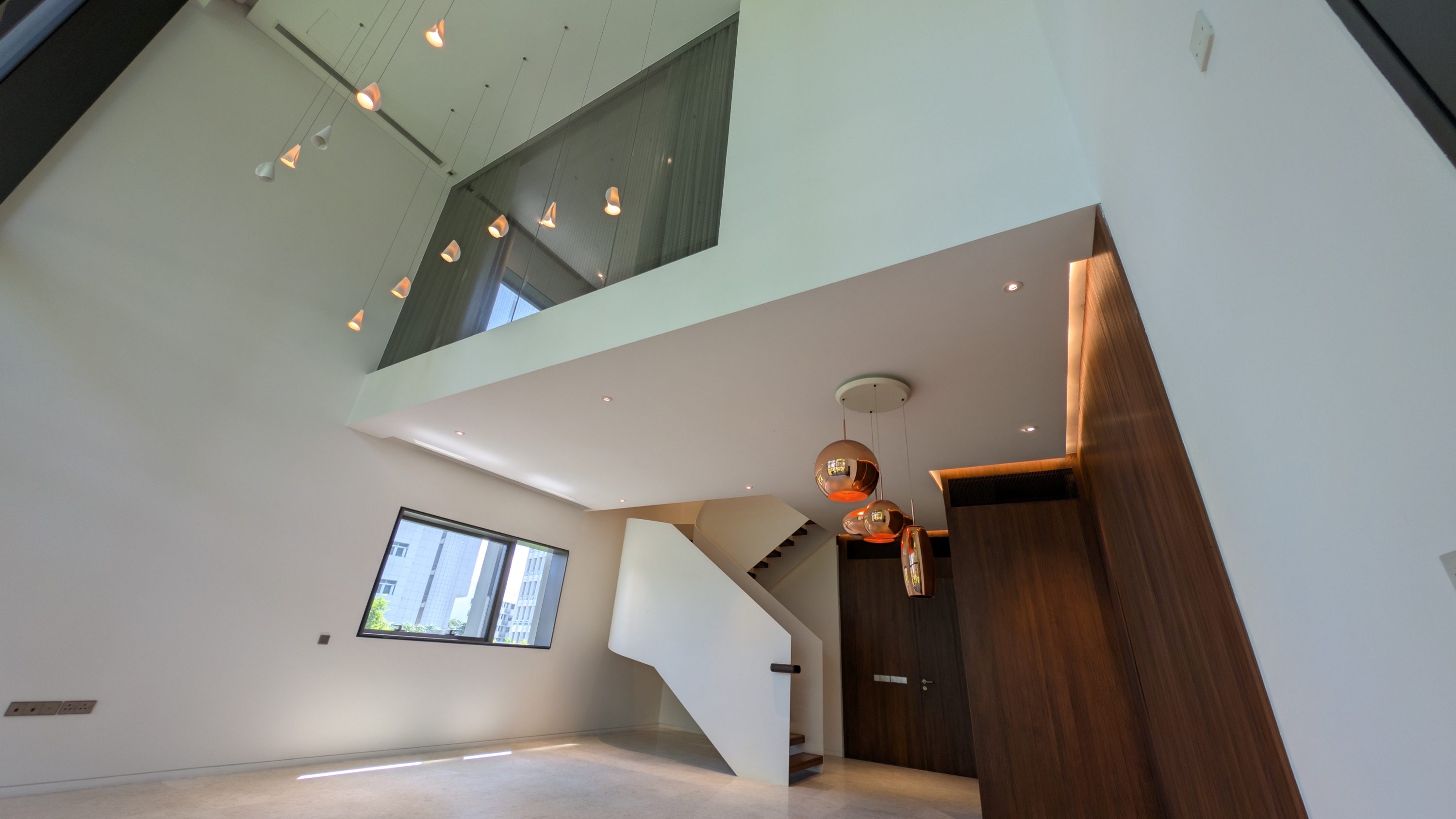 A view of a modern, multi-level living space with high ceilings, featuring a striking light fixture with multiple hanging lamps, a wooden feature wall, and a white staircase leading to an upper landing.
